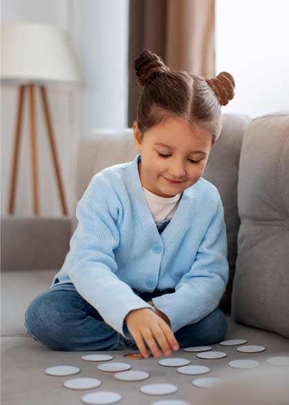A imagem mostra uma menina pequena, aparentando ter entre 4 e 6 anos, sentada no chão com as pernas cruzadas. Ela tem pele clara, cabelos castanhos claros presos em dois pequenos coques no topo da cabeça, e está olhando para baixo com uma expressão concentrada.
Ela veste um cardigã azul claro de tricô sobre uma camiseta branca e calças jeans azuis. Suas mãos estão estendidas à sua frente, interagindo com diversos objetos circulares e planos espalhados pelo chão. Estes objetos parecem ser peças de um jogo ou atividade, possivelmente discos de madeira ou papelão com cores ou desenhos não visíveis.