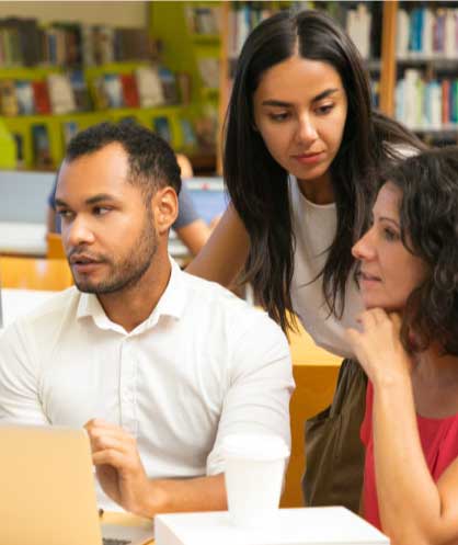 A foto mostra um grupo de três jovens adultos interagindo em um ambiente que parece ser um espaço de estudo colaborativo. Eles estão sentados ao redor de uma mesa de madeira clara, com livros e um laptop espalhados.